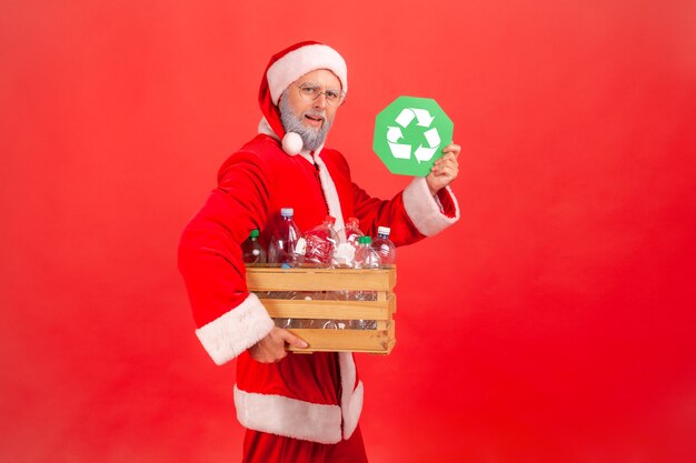 Père noël tenant une boîte avec des bouteilles en plastique et montrant un panneau de recyclage vert à la caméra.