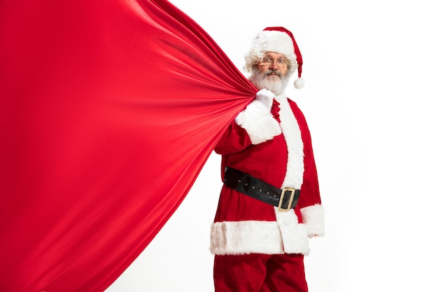 Père Noël tirant un énorme sac plein de cadeaux de Noël isolé sur fond blanc. Modèle masculin caucasien en costume traditionnel. Nouvel An 2020, cadeaux, vacances, ambiance hivernale. Copyspace pour votre annonce.