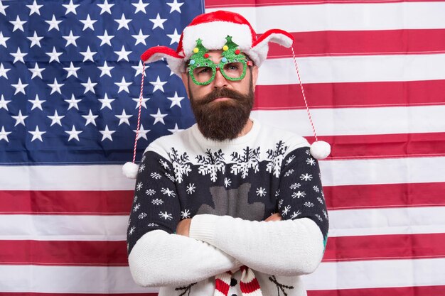 Le Père Noël vous regarde. L'homme barbu porte des lunettes amusantes. Hipster drôle sur fond de drapeau américain. Drôle mais patriotique. Plaisir d'hiver. Joyeux Noël. Idée drôle de costume de nouvel an. Célébration des vacances.