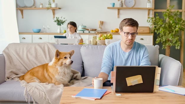 Père occupé travaillant à distance avec un ordinateur portable assis dans la cuisine. Un mignon petit chien est allongé sur le canapé à proximité.