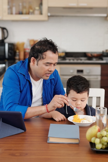 Père parlant à son fils en le nourrissant avec des tranches de fruits pour le déjeuner à la maison