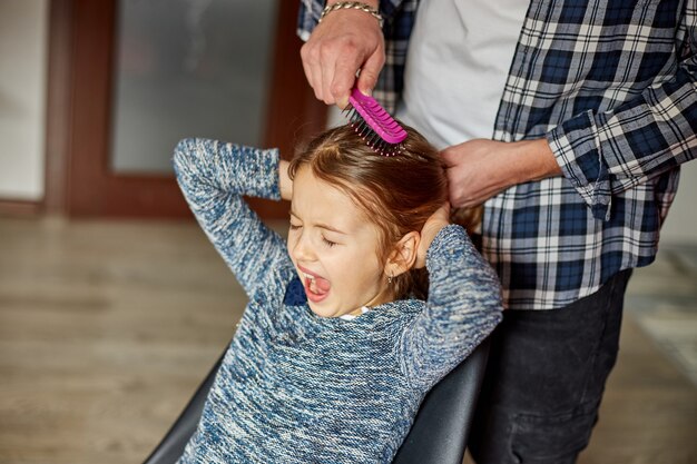 Père peignant, brossant les cheveux de sa fille à la maison, enfant faisant des grimaces sur l'arrachage des cheveux