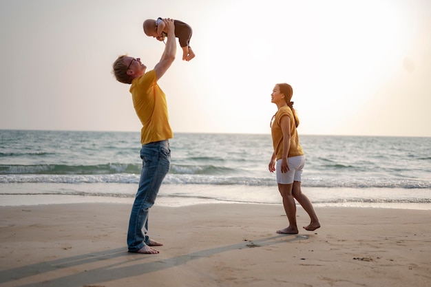 Le Père Porte Et Joue Avec Son Bébé Et Sa Mère S'amusant à La Plage Au Coucher Du Soleil En Vacances. Famille, Plage, Détente, Fils, Bébé, Concept De Style De Vie.