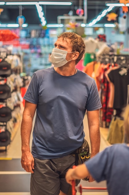 Un père sérieux roule un enfant un garçon dans un chariot de magasin rouge porte un masque bleu protège contre le virus COVID mesure de contrôle de la santé personnelle publique achats en famille pendant la pandémie achat de nouveaux vêtements