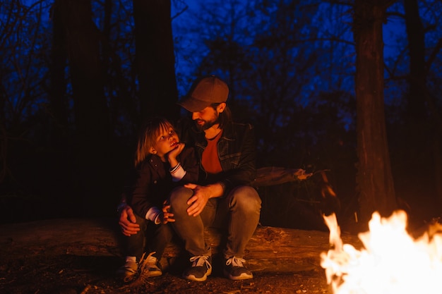 Père et son petit fils assis ensemble sur les bûches devant un feu lors d'une randonnée en forêt la nuit. .