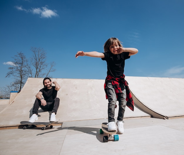 Le père et son petit fils vêtus de vêtements décontractés font du skateboard et s'amusent dans un skate park avec des toboggans à l'extérieur par beau temps.