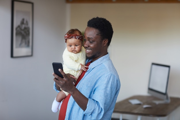 Père souriant africain tenant bébé fille sur ses mains et en utilisant son téléphone portable à la maison