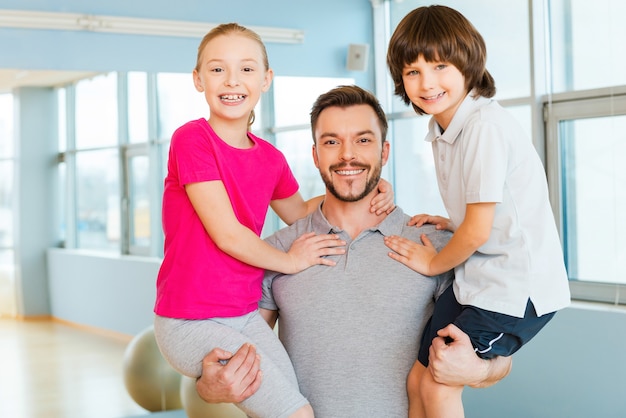 Père sportif avec des enfants. Joyeux jeune père sportif portant ses enfants dans les mains et souriant en se tenant debout dans un club de sport