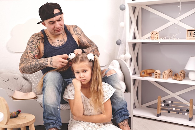 Père tatoué de temps drôle dans une casquette et son enfant jouent à la maison papa fait les cheveux de ses filles...