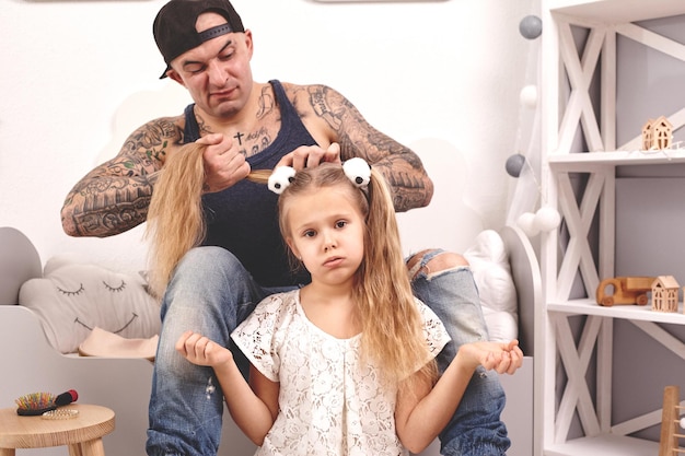 Père tatoué de temps drôle dans une casquette et son enfant jouent à la maison papa fait les cheveux de ses filles...