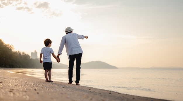 Le père tient la main de son enfant et marche sur la plage au coucher du soleil en vacances en famille