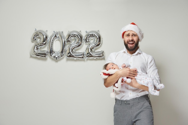 Un père vêtu d'un bonnet de noel en velours rouge tient sa petite fille près de ballons argentés en forme de 2022. Un père serre son petit enfant dans ses bras et chante lors d'une fête du nouvel an.