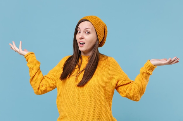 Perplexe jeune femme brune fille en pull jaune et chapeau posant isolé sur fond de mur bleu portrait en studio. Concept de mode de vie des émotions sincères des gens. Maquette de l'espace de copie. Mains écartées.