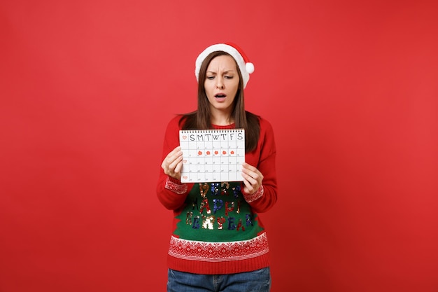 Perplexe Santa girl holding calendrier des périodes féminines pour vérifier les jours de menstruation isolés sur fond rouge. Médical, sanitaire, gynécologique. Bonne année 2019 concept de fête de vacances célébration.