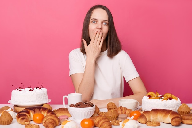 Perplexe surpris femme aux cheveux bruns en t-shirt blanc assis à table avec beaucoup de desserts rompant le régime alimentaire couvrant la bouche avec la paume isolé sur fond rose