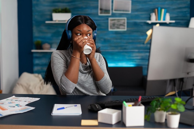 Personne Afro-américaine Souriante Et Utilisant Des écouteurs Pour écouter De La Musique. Femme Portant Un Casque Pour S'amuser Avec Des Chansons Et Regardant Un écran D'ordinateur, Travaillant à Domicile. Divertissement Audio