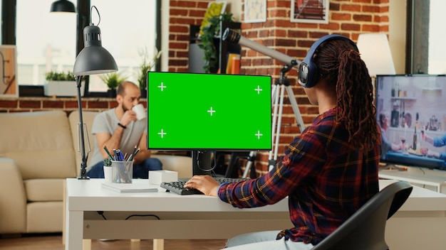 Personne afro-américaine travaillant avec un écran vert et écoutant de la musique sur un casque, utilisant un ordinateur avec affichage à clé chroma et maquette isolée. En regardant un écran vert vierge. Prise de vue au trépied.