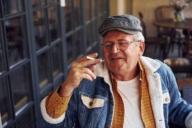 Une personne âgée élégante dans des vêtements à la mode et dans des lunettes est assise dans le café avec une cigarette et des discussions.