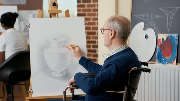 Personne âgée en fauteuil roulant créant un dessin de vase sur toile, participant à un atelier artistique pour développer de nouvelles compétences. Cours d'art pour apprendre à dessiner des croquis d'œuvres d'art professionnelles. Prise de vue à main levée