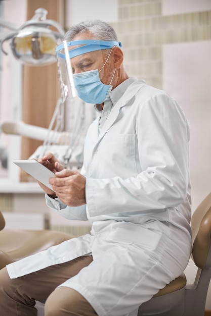 Personne âgée focalisée portant une blouse de laboratoire et un masque chirurgical regardant un écran de tablette tout en travaillant en clinique