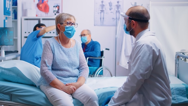 Personne âgée à la retraite lors d'une consultation médicale portant un masque pendant la pandémie de COVID 19. Infirmière à l'arrière avec un vieil homme en fauteuil roulant. Hôpital clinique privé moderne ou établissement de santé.