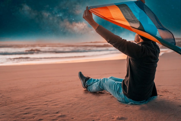 Personne assise sur la plage et tenant un drapeau arc-en-ciel au-dessus de la tête la nuit LGTBQ