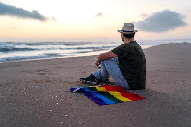 Personne assise sur le rivage de la plage avec drapeau arc-en-ciel regardant le coucher du soleil