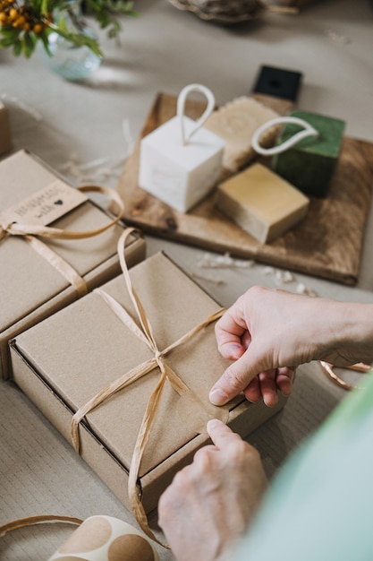Une personne attache un ruban autour de boîtes-cadeaux Kraft sur une table avec des savons faits à la main et des fournitures d'emballage petites