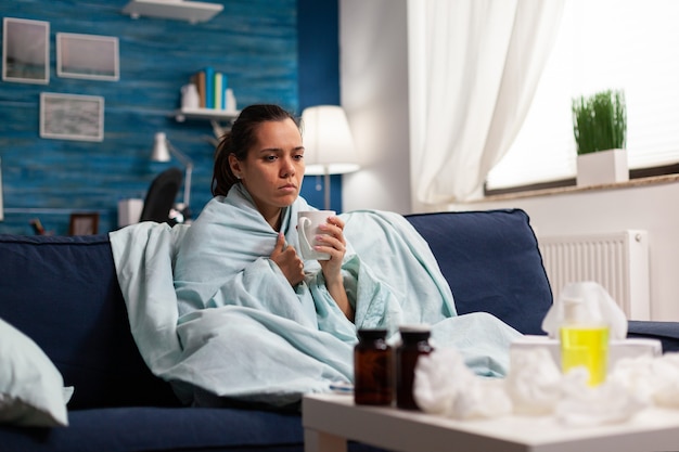 Personne Atteinte D'une Maladie à La Maison Prenant Un Traitement Contre Le Rhume Et La Grippe Assise Sur Un Canapé. Symptômes Saisonniers Et Toux, Tenant Une Tasse Avec Du Thé Ou Un Traitement Médical Contre Les Maladies. Santé De La Gorge