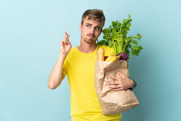 Personne blonde prenant un sac de plats à emporter isolé sur bleu avec les doigts qui se croisent et souhaitant le meilleur