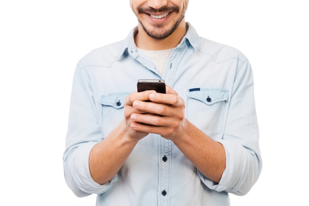 Personne communicative. Photo recadrée d'un beau jeune homme tenant un téléphone portable et souriant en se tenant debout sur fond blanc