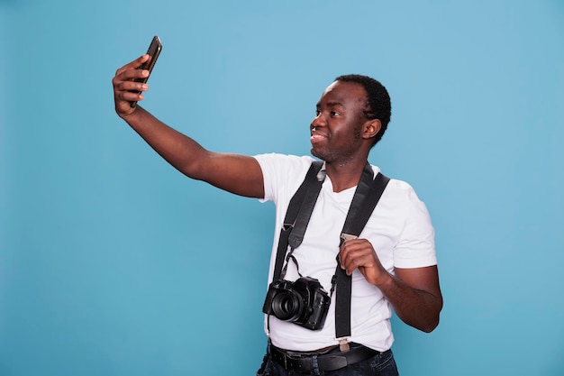 Personne confiante ayant un appareil photo reflex numérique tout en prenant une photo de lui-même avec un téléphone. Photographe professionnel confiant prenant une photo de selfie avec un smartphone tout en se tenant sur fond bleu.
