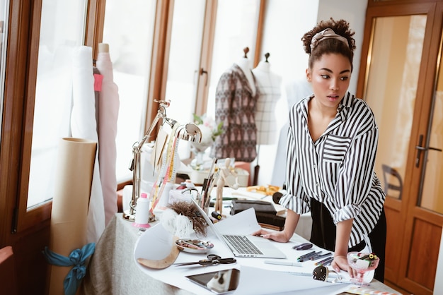 Photo personne créative au studio de design. jeune femme africaine, près de la table de travail avec différents objets d'artisanat. elle se tient près du mannequin de robe