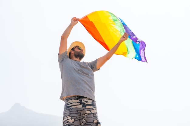 Une personne gay avec un t-shirt gris et un chapeau blanc agitant le drapeau LGBT dans un ciel nuageux