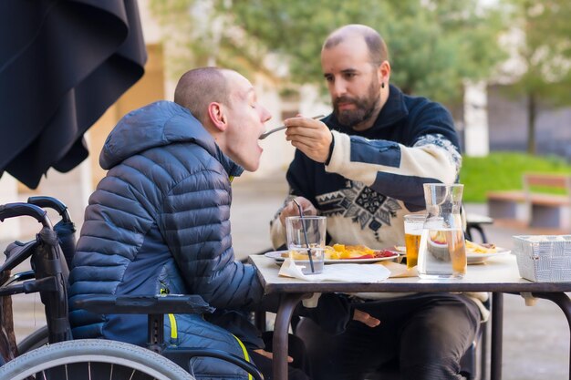 Une personne handicapée mangeant sur la terrasse d'un restaurant et un ami l'aidant à manger