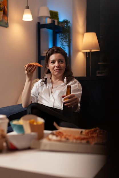 Personne heureuse après le travail en regardant une émission télévisée de sitcom en train de manger une tranche de livraison de pizza chaude assise sur un canapé tenant une bouteille de bière. Femme appréciant le dîner tv à emporter sur le canapé.