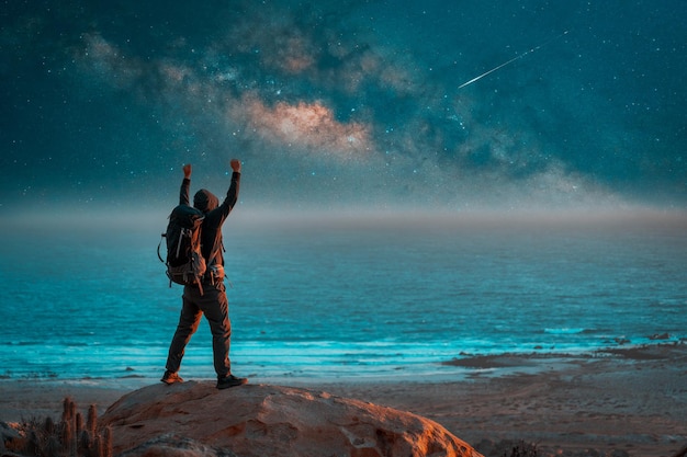 Personne inconnue debout sur le rocher à l'extérieur vue arrière avec les mains levées regardant la voie lactée au-dessus de l'océan