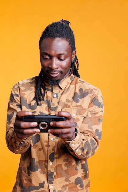 Personne ludique jouant à des jeux vidéo sur téléphone mobile, ayant un tournoi de jeux en ligne. Homme afro-américain appréciant le jeu du cyberespace pendant les loisirs en studio sur fond jaune