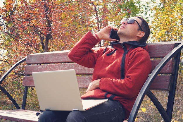 La personne occupée travaille en ligne à l'extérieur du bureau en vacances. jeune homme travaillant sur ordinateur portable dans des vêtements décontractés. Parler sur smartphone, assis dans le parc. résoudre les problèmes par téléphone. Business en ligne.
