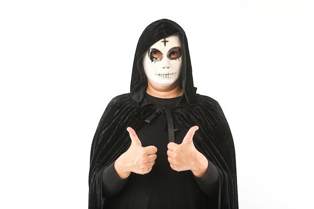 Personne portant un masque blanc avec une croix sur le front et une cape de velours noir avec capuche, levant les pouces vers le haut, sur fond blanc. Concept de carnaval, Halloween et jour des morts.