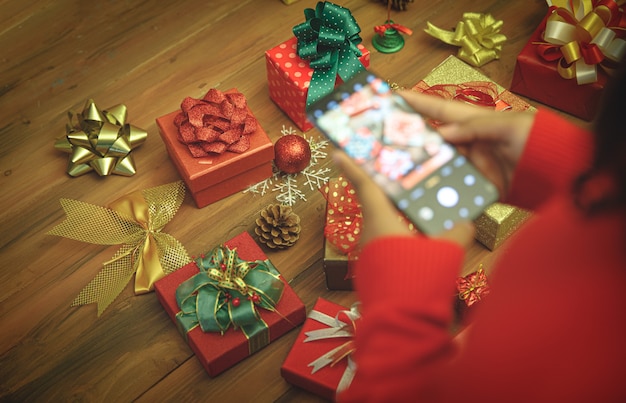 Personne sur un pull utilisant un smartphone pour prendre un instantané et enregistrer une photo d'une luxueuse boîte à cadeaux avec un bel arc coloré et un ornement sur la table, pour mémoriser la charmante décoration de la maison de Noël