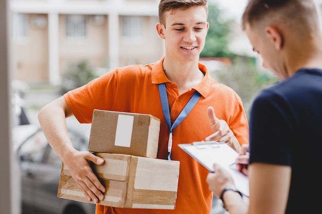 Une personne souriante portant un T-shirt orange et une étiquette nominative livre un colis à un client, qui appose sa signature sur le reçu. Travailleur sympathique, service de livraison de haute qualité.