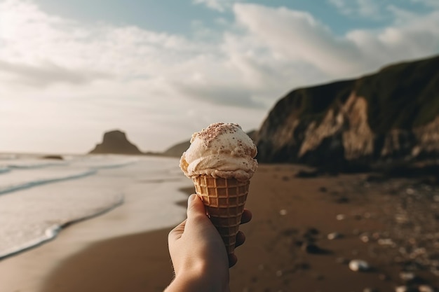 Une personne tenant un cornet de crème glacée sur une plage de sable avec des vagues et des montagnes