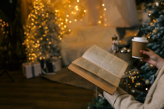 Personne tenant un livre ouvert devant un arbre de Noël décoré avec des lumières brillantes Un cadre confortable avec des tons chauds maisons miniatures et bougies idéales pour les moments de lecture de vacances