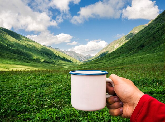 Personne tenant une tasse en émail blanc sur fond de montagnes vertes