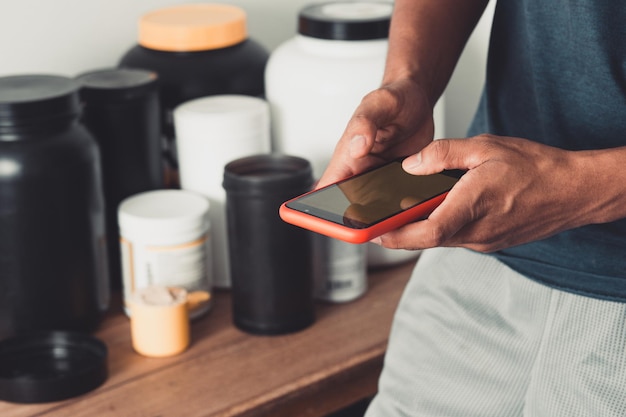 Une personne tient un smartphone à l'aide d'une application de fitness sur fond de conteneurs de suppléments.