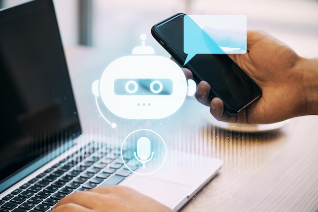 Personne utilisant un smartphone avec une icône de chatbot sur un écran d'ordinateur portable sur un bureau