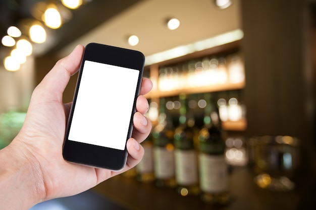 Personne utilisant le support d&#39;écran blanc du smartphone en main avec un fond flou de la barre de whisky pour une fête adulte sur le restaurant.