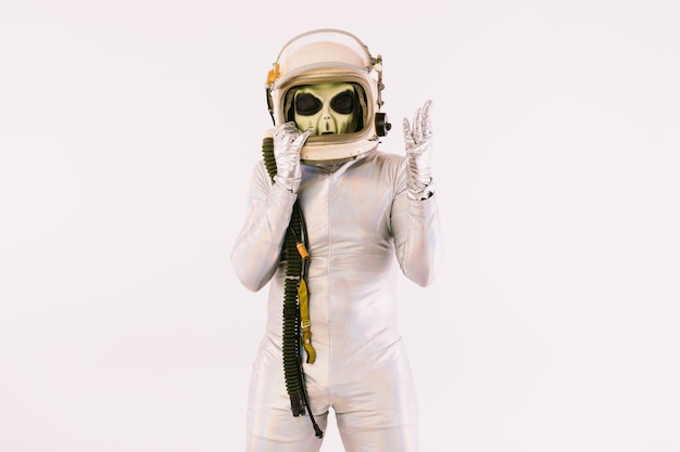 Personne vêtue d'un costume argenté et d'un masque extraterrestre vert, portant un casque de cosmonaute, sur fond blanc