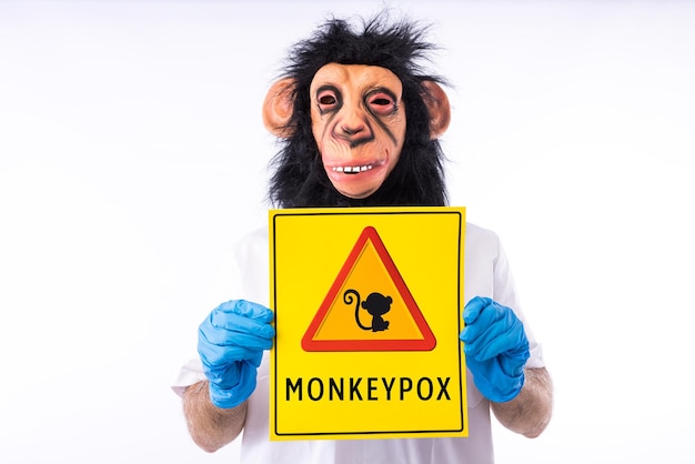 Personne vêtue d'un masque de singe dans un costume d'infirmière médicale tenant une pancarte avec le signe de danger et un singe qui lit MONKEYPOX sur fond blanc Épidémie de virus Nigeria et variole